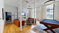 Located at 31 Elm St in Springfield, MA, this thoughtfully designed amenity space offers a generous, open single-floor layout that seamlessly blends comfort, entertainment, and function. The main lounge area is anchored by a dramatic floor-to-ceiling marble-clad fireplace wall, complete with a wall-mounted TV and a glowing linear electric fireplace. Warm hardwood flooring runs throughout, complemented by soaring ceilings with a coffered tile finish and a statement ring pendant light. Tall windows dressed in floor-length patterned curtains flood the space with natural light and offer views of the surrounding streetscape. A plush seating arrangement — including a rich burnt-orange velvet sofa and sculptural upholstered chairs — creates an inviting social atmosphere, while a full-size billiards table adds a recreational focal point. A dedicated meeting area features a sleek white conference table, cognac leather chairs, and its own window with outdoor views, accented by a hexagonal-patterned ceiling. The kitchen is fitted with white cabinetry, a striking navy hexagonal tile backsplash, stainless steel appliances, and a large island with pendant lighting above — ideal for catering or community gatherings. Together, these areas form a vibrant, well-appointed communal destination.