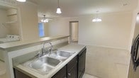 This single-floor space showcases an open-concept design that connects the kitchen and living areas into one cohesive layout. The kitchen features a double-basin stainless steel sink set into light-toned countertops, complemented by dark lower cabinetry and a built-in dishwasher. A pendant light fixture hangs directly above the sink area, providing focused task lighting. A pass-through counter with a low partition wall opens the kitchen to the adjacent living space, enhancing the sense of openness and allowing natural light from a window in the living area to filter through. The living area is finished with neutral carpet flooring and is illuminated by an elegant chandelier, adding a decorative touch overhead. A ceiling fan in the living space provides additional air circulation. Crown molding runs along the upper perimeter of the walls, lending a refined architectural detail throughout. The neutral wall tones and light fixtures create a bright, clean ambiance. A doorway visible in the background suggests additional rooms beyond, contributing to the overall functionality of the floor plan.