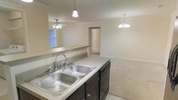 This single-floor space showcases an open-concept design that connects the kitchen and living areas into one cohesive layout. The kitchen features a double-basin stainless steel sink set into light-toned countertops, complemented by dark lower cabinetry and a built-in dishwasher. A pendant light fixture hangs directly above the sink area, providing focused task lighting. A pass-through counter with a low partition wall opens the kitchen to the adjacent living space, enhancing the sense of openness and allowing natural light from a window in the living area to filter through. The living area is finished with neutral carpet flooring and is illuminated by an elegant chandelier, adding a decorative touch overhead. A ceiling fan in the living space provides additional air circulation. Crown molding runs along the upper perimeter of the walls, lending a refined architectural detail throughout. The neutral wall tones and light fixtures create a bright, clean ambiance. A doorway visible in the background suggests additional rooms beyond, contributing to the overall functionality of the floor plan.