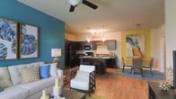 This single-floor residence showcases an open-concept layout that integrates the living, dining, and kitchen areas into one cohesive, flowing space. The living area is anchored by a ceiling fan, warm hardwood-style flooring, and a striking teal accent wall that adds a bold pop of color, complemented by a cheerful yellow accent wall near the dining space. Natural light fills the interior, enhancing the bright and welcoming ambiance throughout. The kitchen is well-appointed with dark espresso cabinetry, black appliances including a refrigerator, microwave, and range, and a granite-topped island that doubles as a breakfast bar with pendant lighting overhead. Track lighting adds additional illumination to the kitchen workspace. The dining area, positioned adjacent to the kitchen, offers an intimate setting ideal for everyday meals or entertaining. The living area provides ample room for comfortable seating arrangements, with decorative touches that lend personality to the space. The consistent hardwood-style flooring throughout ties the open layout together seamlessly, creating a unified and stylish living environment that is both functional and visually dynamic.
