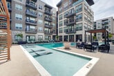 Located at 9305 Montview Blvd in Aurora, Colorado, this outdoor amenity deck offers a thoughtfully designed resort-style experience set against the backdrop of a modern multi-story residential building. The centerpiece is a generously sized swimming pool with crystal-clear water, complemented by in-pool lounge chairs and a separate spa, ideal for relaxation. The surrounding pool deck is finished in clean concrete and lined with comfortable seating areas, including lounge chairs and shaded pergola structures that provide respite from the Colorado sun. A standout feature is an outdoor entertainment wall equipped with a large-screen television and a built-in linear fireplace, creating a functional and inviting gathering space. Glass panel railings define the perimeter, maintaining unobstructed views of the open surrounding landscape, while string lights strung between posts add a warm, ambient atmosphere for evening use. Raised brick-bordered planters with trees and native landscaping add natural texture throughout the deck. The building's multi-story facade, featuring private balconies and large windows, frames the courtyard on multiple sides. Additional outdoor seating zones with modern furniture provide versatile spaces for socializing or quiet enjoyment.
