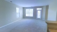 This single-floor living space features a generously sized main room finished with soft, neutral-toned wall-to-wall carpeting that adds warmth underfoot. The room is bathed in natural light, courtesy of multiple windows fitted with white horizontal blinds, including a large central window and a secondary window along the adjacent wall, both framing views of surrounding greenery. A glass-panel exterior door further enhances the sense of openness, allowing additional daylight to filter in while providing convenient access to the outdoors. The walls are painted in a calming light gray-beige tone, creating a clean, neutral backdrop well-suited to a variety of décor styles. White baseboards offer a crisp contrast to the soft flooring and walls, contributing to a tidy, polished finish throughout. The layout is open and unobstructed, making the space highly versatile and accommodating for various furniture arrangements. The overall ambiance is light, airy, and welcoming, with a straightforward design that emphasizes functionality and comfort. The abundance of natural light and the view of outdoor foliage through the windows add a refreshing, connected-to-nature quality to the interior.