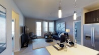 This thoughtfully designed single-floor residence offers a seamless blend of modern aesthetics and functional living. The open-concept layout connects the dining and living areas, creating a spacious, airy atmosphere ideal for both everyday living and entertaining.

The living area is anchored by a deep navy sectional sofa, complemented by a sleek coffee table and a cozy accent chair. A large piece of wall art adds a sophisticated focal point, while hardwood-style flooring runs throughout, lending warmth and continuity to the space. Large windows and glass-paneled doors flood the room with natural light, offering a bright and welcoming ambiance.

Overhead, modern cylindrical pendant lights hang elegantly, providing both task and ambient illumination. The dining area, positioned adjacent to the living space, features a clean-lined table set with stylish place settings, enhanced by additional pendant lighting that creates an intimate yet refined dining experience.

The cool, neutral color palette — composed of soft grays, deep blues, and crisp whites — gives the space a calm and contemporary character. The thoughtful arrangement of furnishings and lighting ensures a cohesive, polished look throughout, making this a comfortable and visually compelling single-floor residence.