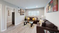 This single-floor residence offers a cohesive and modern living experience with a clean, contemporary aesthetic carried throughout every room. Light gray walls and light-toned hardwood-style flooring unify the space, creating a bright and welcoming atmosphere.

The living area serves as the heart of the home, accommodating generous seating with two dark brown sofas accented by yellow and neutral throw pillows. Recessed lighting and a large window with horizontal blinds allow natural light to fill the room, while a glass-top coffee table and colorful abstract wall art add character and style. The space opens to a hallway that connects the remaining rooms seamlessly.

The kitchen is efficiently designed with crisp white shaker cabinetry extending to the ceiling, offering ample storage. Stainless steel appliances — including a refrigerator, range, and dishwasher — complement the clean white countertops. A window above the sink provides natural light, making the kitchen feel open and functional.

Both bedrooms are well-proportioned and share a consistent design language, each featuring light hardwood-style flooring, ceiling fans with integrated lighting, and windows fitted with blinds that allow for adjustable natural light. The bedrooms provide a calm, restful environment with neutral wall tones.

The bathroom is neatly appointed with a white vanity, a tub-shower combination, and tiled flooring, maintaining the home's clean and practical design throughout.