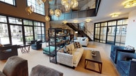 This two-story amenity space blends industrial-modern design with upscale comfort across two distinct levels. The main floor commands immediate attention with its soaring double-height ceilings and dramatic floor-to-ceiling black-framed windows that flood the space with natural light and offer views to the outdoors. A cascading cluster of sculptural globe pendant lights serves as a striking focal centerpiece overhead. Multiple seating arrangements — featuring plush sofas, accent chairs, and curated coffee tables — are organized across the main floor to encourage both social gathering and quiet relaxation. A large decorative wall mural adds artistic depth, while a media console with a wall-mounted TV anchors one end of the lounge. A cozy secondary seating nook with a fireplace, wall art, and bistro-style seating provides an intimate retreat off the main area. An industrial-style staircase with cable railings connects to the upper level, where a dedicated games floor awaits. This second level offers a foosball table, ping-pong table, billiards table, and a bar-height counter lined with red stools beneath wall-mounted screens — ideal for entertainment and social play. Textured tile-pattern carpet, artistic wall murals, and steel-framed glass partitions throughout lend the space a refined yet dynamic character.
