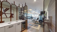 This single-floor fitness center near Westerly Creek Trail in Denver's 80238 neighborhood offers a generously sized, thoughtfully designed space for comprehensive workouts. The layout is divided into distinct functional zones, providing dedicated areas for both cardiovascular training and strength work.

The cardio zone features a row of treadmills positioned near large windows and glass doors that allow natural light to fill the space and provide views to the outdoors. Ceiling fans and distinctive cloud-shaped light fixtures are mounted on a coffered ceiling, contributing to an airy, comfortable atmosphere during exercise.

The strength training area is well-appointed with multi-rack systems, extensive dumbbell arrays, adjustable benches, cable machines, and functional training equipment. Rubber flooring underlays the free weight zones, while a bold multi-toned modular carpet in gray, tan, and orange geometric patterns covers the remaining floor area, adding visual interest throughout.

Near the entrance, arched full-length mirrors framed in warm wood tones serve as a stylish room divider, complemented by a refreshment station with white cabinetry and a built-in beverage cooler. A terracotta-tiled accent wall further enhances the modern, design-forward aesthetic of this fully equipped fitness facility.