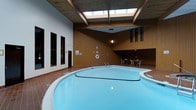 This indoor aquatic facility centers around a spacious, kidney-shaped swimming pool with calm, clear blue water. The pool is equipped with metal entry railings and a life ring mounted nearby, reflecting a strong emphasis on safety. The surrounding pool deck is finished in warm terracotta tile, providing a slip-resistant and visually inviting border around the water.

The architecture of the space is defined by its striking wood-paneled walls in a rich brown tone, giving the interior a warm and welcoming ambiance. The ceiling draws the eye upward, where a large skylight allows natural light to pour into the space, enhancing the brightness of the water and reducing the enclosed feeling common to indoor pools. Structural beams add to the architectural character of the ceiling design.

Along one wall, a row of dark-framed doorways leads to what appear to be changing or utility rooms, providing functional access for users of the facility. On the opposite side, a casual seating area features a round table and chairs, offering a comfortable spot for relaxation or supervision. Wall-mounted lighting complements the natural light from above, ensuring the space remains well-lit throughout the day. Overall, this facility combines practical amenities with a thoughtfully designed interior for a comfortable aquatic experience.