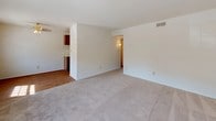 This single-floor residence offers a practical and straightforward layout with two bedrooms and one and a half bathrooms. The living room is a generous, open space with neutral-toned carpet, white walls, and a window that allows natural light to brighten the room. It connects seamlessly to the kitchen area, which features wood-look flooring, dark wood upper cabinetry, light countertops, and includes appliances such as an electric range and dishwasher. A half-wall partition neatly defines the boundary between the kitchen and living areas. Both bedrooms are comfortably sized, finished with the same neutral carpet, and feature clean white walls, offering a blank canvas for personalization. The full bathroom is outfitted with warm beige tile surrounding a full-size soaking tub and shower combo, a wall-mounted mirror, and classic white fixtures. The overall color palette throughout the unit is light and neutral, creating a clean, move-in-ready atmosphere. The floor plan, as seen in the overhead view, reveals a well-organized and efficient use of space, with clearly defined rooms and good connectivity between living, sleeping, and utility areas.