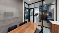 This conference room delivers a refined, contemporary aesthetic through a thoughtful combination of materials and design elements. The centerpiece is a solid wood conference table paired with black ergonomic chairs, providing comfortable seating for multiple attendees. One wall features a striking concrete texture finish, lending an industrial character to the space, and is adorned with two framed geometric art pieces accented by a brass decorative floor lamp. A wall-mounted flat-screen display is positioned on the opposite wall for presentations and video conferencing needs.

A glass whiteboard panel mounted to the concrete wall provides a functional writing surface without disrupting the room's visual cohesion. Floor-to-ceiling black-framed glass partitions define the room's boundaries while maintaining an open, transparent connection to the surrounding office environment. The patterned tile flooring adds a subtle decorative layer underfoot. Adjacent to the room's glass entry, a compact wooden credenza with a marble top and a secondary wall-mounted display provide additional utility. Recessed lighting ensures the space is well-lit, contributing to a professional and polished atmosphere suited for focused meetings and collaborative work sessions.