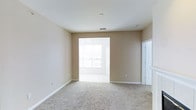 This single-floor space presents a clean, open layout designed for comfortable everyday living. The main living area features wall-to-wall carpet in a neutral beige tone, complementing the warm, tan-painted walls and crisp white trim and baseboards throughout. The room offers a generous sense of space, with standard ceilings fitted with a central ventilation duct, keeping the area well-aired.

A wide open doorway connects the carpeted living area to an adjacent room, which features tile or hard flooring and is bathed in abundant natural light from large windows. A ceiling fan is visible in this adjoining space, adding to its functionality and comfort. The bright light filtering through creates an inviting, airy atmosphere that carries into the main living area.

To the right, a white paneled door with decorative molding detail provides access to an additional room or entryway, adding architectural interest to the otherwise clean-lined design. The overall aesthetic is understated and neutral, offering a versatile backdrop well-suited for a variety of furnishing styles and personal preferences.