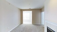 This single-floor space presents a clean, open layout designed for comfortable everyday living. The main living area features wall-to-wall carpet in a neutral beige tone, complementing the warm, tan-painted walls and crisp white trim and baseboards throughout. The room offers a generous sense of space, with standard ceilings fitted with a central ventilation duct, keeping the area well-aired.

A wide open doorway connects the carpeted living area to an adjacent room, which features tile or hard flooring and is bathed in abundant natural light from large windows. A ceiling fan is visible in this adjoining space, adding to its functionality and comfort. The bright light filtering through creates an inviting, airy atmosphere that carries into the main living area.

To the right, a white paneled door with decorative molding detail provides access to an additional room or entryway, adding architectural interest to the otherwise clean-lined design. The overall aesthetic is understated and neutral, offering a versatile backdrop well-suited for a variety of furnishing styles and personal preferences.