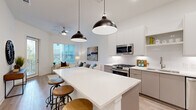 Located at 652 Angier Ave NE in Atlanta's vibrant 30308 zip code, this single-floor residence showcases a contemporary design with clean lines, light-toned hardwood-style flooring, and a bright, neutral color palette throughout. The open-concept living and kitchen area serves as the heart of the home, where a generous island with white quartz countertops and industrial-style pendant lighting anchors the space. The kitchen is outfitted with two-tone cabinetry—white uppers and warm gray lowers—paired with a full suite of stainless steel appliances including a gas range, built-in microwave, dishwasher, and French door refrigerator, all complemented by a classic white tile backsplash. The living area benefits from large windows and a glass-paneled exterior door that flood the space with natural light and offer outdoor views. The bedroom is a serene retreat featuring elevated ceilings, a ceiling fan, and a bright window, with direct access to a spacious walk-in closet outfitted with built-in shelving and double hanging rods. The bathroom presents a frameless glass walk-in shower with large-format tile surround, a built-in niche, open shelving, and wood-look flooring, combining functionality with modern elegance.