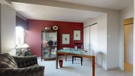This single-floor residence offers a well-appointed layout with a cohesive design aesthetic defined by rich red accents, neutral tones, and plush carpeting throughout. The living room is a standout space, featuring an expansive bank of large windows that frame sweeping panoramic city and river views, filling the room with natural light. Comfortable seating surrounds a dark wood coffee table, complemented by a substantial entertainment armoire.

The dining area accommodates an elegant glass-top table beneath a contemporary pendant light fixture, with framed artwork adorning the walls. Adjacent to the dining space, a dedicated study or den features a bold crimson accent wall, a glass-top desk, and a curved glass display cabinet, creating a sophisticated and functional workspace.

The kitchen is efficiently designed with white cabinetry, light countertops, and a full suite of appliances including a gas range and dishwasher. A carpeted hallway connects the private quarters, leading to well-sized bedrooms, each featuring soft carpeting, closet storage, and windows with city views. The primary bedroom includes rich drapery and dark wood furniture, reinforcing the home's consistent, polished design throughout.