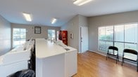 This single-floor, self-serve laundry facility is designed to offer residents a convenient and functional space for their laundry needs. The room features warm wood-look flooring throughout, complemented by neutral gray walls and clean white trim that give the space a tidy, welcoming atmosphere. Vaulted ceiling sections add an open, airy quality to portions of the room, while flush-mounted fluorescent lighting ensures the space is well-lit at all hours.

A bank of stacked commercial dryers lines one wall, providing ample drying capacity. Alongside these, a row of top-load washing machines runs parallel to a central white folding counter, offering generous surface area for sorting and folding laundry. A dedicated folding table with modern hairpin legs provides additional workspace near the windows.

Natural light filters in through multiple windows fitted with horizontal blinds, offering views of surrounding greenery. A wall-mounted clock and a "Laundry – Open 24 Hours / Self-Serve" sign reinforce the facility's around-the-clock availability. A Coca-Cola vending machine adds a practical amenity for users. The space is accessed through a keyed entry door, ensuring security for residents. Overall, this laundry facility is well-equipped and efficiently laid out to serve a residential community.