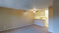 This single-floor residential space features a practical and functional layout designed for comfortable everyday living. The main living area is open and airy, finished with neutral-toned walls and carpeted flooring that adds warmth underfoot. A ceiling fan with a built-in light fixture is centrally mounted beneath a textured popcorn ceiling, providing both air circulation and ambient lighting to the room. Clean white baseboards frame the perimeter of the space, adding a crisp finishing touch. The living area flows seamlessly into the kitchen, which is accessible via a raised breakfast bar counter that doubles as a casual dining or prep surface. The kitchen is equipped with white cabinetry offering ample storage, a sink positioned beneath upper cabinets, and standard appliances. The pass-through design of the kitchen keeps the space connected to the main living area, making it ideal for entertaining or multitasking. The overall color palette is warm and understated, with tan and beige tones creating a cohesive and inviting atmosphere throughout. The single-floor layout ensures ease of movement and accessibility across all areas of the space.