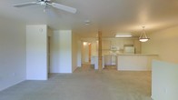 This single-floor space features an open-concept layout that seamlessly connects the living area to the kitchen. The living area is finished with neutral wall-to-wall carpeting, creating a warm and understated foundation for the room. A ceiling fan is mounted overhead, providing both functional air circulation and a subtle design element.

A distinctive natural wood post serves as a structural and visual centerpiece, anchoring the transition between the living space and the kitchen zone. The kitchen is outfitted with white cabinetry, a white electric range, a built-in microwave, and a refrigerator, all arranged in an efficient layout. A peninsula counter extends into the open area, offering additional workspace and a casual dining or breakfast bar option. Fluorescent lighting above the kitchen provides consistent illumination throughout the cooking area.

Adjacent to the main open area, a hallway leads further into the unit, with doorways visible that suggest additional rooms beyond. The overall ambiance is clean, bright, and functional, with neutral tones throughout lending the space a versatile and move-in-ready character.