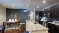 This single-floor space showcases an open-concept layout that connects the kitchen and dining areas in a cohesive, contemporary design. The kitchen is a standout feature, centered around a large granite-topped island with a stainless steel undermount sink and bar seating. Two pendant lights hang above the island, adding both functional illumination and a decorative touch. Dark espresso cabinetry lines the kitchen walls, complemented by a teal subway tile backsplash, stainless steel appliances — including a French door refrigerator, microwave, and range — and ample upper and lower storage. Recessed lighting throughout ensures the space remains bright and well-lit. The dining area sits adjacent to the kitchen, featuring a wooden dining table with chairs set atop a patterned area rug over rich hardwood flooring. A bold, deep teal-gray accent wall provides a dramatic backdrop, adorned with a large decorative wall installation that adds an artistic focal point. The overall ambiance is modern and sophisticated, with a warm undertone created by the combination of dark cabinetry, natural wood tones, and soft lighting.