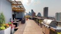 Located in the heart of Seattle's Belltown neighborhood, this rooftop deck at 206 Bell St offers a dynamic outdoor retreat with sweeping panoramic views of the Seattle skyline and Elliott Bay waterfront. The deck is finished with warm wood tile flooring and anchored by a covered lounge area featuring a bold yellow pergola structure and corrugated metal siding, lending the space a distinctive industrial-modern aesthetic. Beneath the pergola, a central fire pit table is surrounded by plush navy blue cushioned sofas and lounge chairs, with a mounted flat-screen TV overhead — ideal for gatherings in any season. Adjacent to the lounge, a bar-height communal dining area with sleek black tables and stools runs along the perimeter railing, offering unobstructed skyline views. A stainless steel gas grill station provides a well-equipped outdoor cooking setup. Vibrant planter boxes filled with ornamental grasses and colorful flowers add greenery throughout, softening the urban surroundings. A narrow interior corridor lined with corrugated metal walls connects rooftop amenities and includes individual storage units. The space combines functional amenities with stunning elevated views, creating an exceptional urban outdoor environment.
