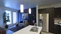 This single-floor space showcases a thoughtfully designed open-concept layout that seamlessly connects the kitchen and living areas. The kitchen features a clean white island that serves as both a workspace and casual gathering point, complemented by dark upper cabinetry and a full-size stainless steel French door refrigerator with a built-in water dispenser. Two cylindrical pendant lights hang overhead, providing focused task lighting while adding a contemporary aesthetic touch.

The living area is anchored by a striking deep navy accent wall that creates a bold visual backdrop and lends a sense of depth and sophistication to the space. A gray tufted sofa and a classic leather armchair offer comfortable seating, while a patterned area rug defines the lounge zone. Framed artwork and small gallery-style frames adorn the accent wall, adding personality. A window fitted with blinds allows natural light to filter into the space. The light wood-tone flooring runs throughout, tying the open-plan areas together cohesively and enhancing the overall modern, urban ambiance of the unit.