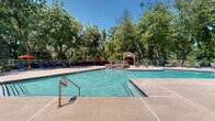 Located at 500 Maxham Rd in Austell, Georgia, this single-story community facility is thoughtfully designed to offer a resort-style outdoor experience. The centerpiece of the property is a generously sized swimming pool with clear turquoise water, accessible via entry steps with metal handrails. The pool's geometric shape includes a shallow wading area, making it suitable for a range of users. A charming wooden gazebo with a pitched roof stands adjacent to the pool, complemented by a stone accent wall and decorative potted plants that enhance the natural aesthetic. The expansive concrete pool deck provides ample space for lounging, furnished with multiple reclining chaise lounges and an umbrella-shaded seating and dining area — ideal for outdoor gatherings. The clubhouse building features light yellow horizontal siding, a pitched roofline with a circular accent window, and distinctive red double-door entries. Large windows along the building's facade allow natural light to flow through while offering views of the surrounding landscape. The entire facility is enveloped by a dense canopy of mature trees, creating a shaded, park-like atmosphere. A metal railing fence secures the perimeter of the pool area, ensuring safety while preserving the open, airy feel of this well-appointed community amenity.