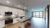 This contemporary two-floor residence at 3700 National Dr in Raleigh, NC offers a well-appointed living experience defined by modern design and quality finishes. The open-concept kitchen serves as a central highlight, featuring crisp white cabinetry extending to the ceiling, a herringbone-patterned tile backsplash, dark quartz countertops, a large center island with pendant lighting, and a full suite of stainless steel appliances including a refrigerator, range, microwave, dishwasher, and a built-in wine cooler. Warm wood-look flooring flows throughout the main living areas, complemented by large windows that invite abundant natural light and views of the surrounding neighborhood. The bedrooms are carpeted for comfort, with one featuring floor-to-ceiling windows and a ceiling fan, creating a bright and airy retreat. The primary bathroom is finished with large-format marble-style wall tiles, a frameless glass walk-in shower with brushed gold fixtures, and a well-lit vanity. Storage is thoughtfully addressed through organized walk-in closets with built-in shelving, hanging rods, and drawer units. Additional bedrooms are generously sized with carpeted floors and easy access to shared spaces. Throughout, neutral tones, gray trim, and recessed lighting create a cohesive, modern aesthetic across both floors.