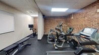 This 560 square foot fitness facility, located at 239 E Boundary Ave in York, PA, offers a well-equipped space for exercise enthusiasts. The single-floor layout maximizes functionality within its compact footprint. Exposed brick walls throughout lend a rustic, industrial aesthetic, contrasting with the modern exercise equipment.

The facility features distinct areas for various workout routines. A cardio section houses treadmills, elliptical machines, and stationary bikes, all equipped with digital displays for tracking progress. The strength training area includes a rack of free weights and resistance machines for targeted muscle group exercises.

Overhead, recessed lighting and skylights provide ample illumination, creating a bright and energizing atmosphere. The dark, rubberized flooring offers durability and shock absorption, ideal for high-impact activities. Climate control systems ensure comfort during intense workouts.

Thoughtful design elements include strategically placed mirrors, water bottle filling stations, and storage solutions for personal belongings. The layout promotes easy navigation between equipment, allowing for efficient workout transitions. This well-appointed gym space combines functionality with an inviting ambiance, catering to a range of fitness needs within its limited square footage.