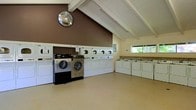 This single-story shared laundry facility is thoughtfully designed to accommodate high-volume use with a well-organized, open floor plan. The room's most striking architectural feature is its vaulted ceiling with exposed white-painted wooden beams, which adds a sense of height and airiness to the space. A bold brown accent wall serves as a visual anchor, complementing the light-toned walls and neutral-colored flooring throughout.

The facility is equipped with an extensive lineup of top-load washing machines arranged along one wall, paired with a row of stacked front-load dryers positioned nearby. Additionally, a set of high-capacity, commercial-grade front-load washing machines is prominently featured, catering to larger laundry needs. A folding counter runs along one side of the room, providing a practical surface for sorting and folding laundry.

Natural light filters into the space through a window overlooking greenery, contributing to a pleasant and welcoming atmosphere. A convex safety mirror is mounted on the wall near the ceiling, enhancing visibility throughout the room. The open layout ensures ample floor space for comfortable movement between machines.