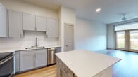 This single-floor residence showcases a thoughtfully designed open-concept layout with warm hardwood-style flooring throughout. The kitchen is a standout feature, equipped with gray shaker-style cabinetry, a herringbone tile backsplash, white quartz countertops, a central island, and a full suite of stainless steel appliances including a refrigerator, range, microwave, and dishwasher. Recessed lighting illuminates the space, which flows seamlessly into the living area. The living room is bright and airy, featuring French doors that open to an outdoor balcony, a ceiling fan, and ample natural light. A built-in desk nook offers a dedicated workspace conveniently positioned near the kitchen. The bedroom benefits from a large window framing lush greenery views, complemented by clean neutral walls and consistent hardwood-style flooring. The bathroom features a wide dual-sink vanity with blue-gray cabinetry, white quartz countertops, a large mirror, and wood-look flooring. Throughout, the space maintains a clean, contemporary aesthetic with neutral tones, modern finishes, and a cohesive design sensibility.