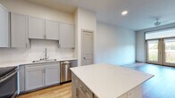 This single-floor residence showcases a thoughtfully designed open-concept layout with warm hardwood-style flooring throughout. The kitchen is a standout feature, equipped with gray shaker-style cabinetry, a herringbone tile backsplash, white quartz countertops, a central island, and a full suite of stainless steel appliances including a refrigerator, range, microwave, and dishwasher. Recessed lighting illuminates the space, which flows seamlessly into the living area. The living room is bright and airy, featuring French doors that open to an outdoor balcony, a ceiling fan, and ample natural light. A built-in desk nook offers a dedicated workspace conveniently positioned near the kitchen. The bedroom benefits from a large window framing lush greenery views, complemented by clean neutral walls and consistent hardwood-style flooring. The bathroom features a wide dual-sink vanity with blue-gray cabinetry, white quartz countertops, a large mirror, and wood-look flooring. Throughout, the space maintains a clean, contemporary aesthetic with neutral tones, modern finishes, and a cohesive design sensibility.
