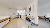 Located at 569 Lexington Avenue in Midtown Manhattan, this single-bedroom unit offers a thoughtfully organized layout that maximizes every inch of its compact footprint. The interior design follows a clean, minimalist aesthetic, with bright white walls, light wood-tone flooring, and recessed lighting that collectively create a bright and open ambiance throughout the space.

The main living area integrates a dedicated workspace, featuring a natural wood-top desk paired with an ergonomic task chair, positioned to take advantage of natural light from a wide bank of windows fitted with roller shades. These windows frame views of the surrounding Midtown streetscape and allow generous daylight to fill the room.

The sleeping area accommodates a lofted-style single bed with built-in under-bed drawer storage, alongside a wardrobe unit, making efficient use of vertical space. A floor lamp provides warm supplemental lighting in this area.

The kitchenette is neatly tucked into one corner of the unit, featuring white cabinetry, a compact countertop with sink, microwave, and a mini-refrigerator — providing all essential cooking and dining conveniences. A separate bathroom with marble-look wall finishes and a window is accessible from the main living area, completing the unit's functional layout.