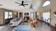 This thoughtfully designed single-floor space makes an immediate impression with its dramatic vaulted ceilings that rise to striking heights, creating an open and airy atmosphere throughout. A bold teal accent wall with geometric angles adds a vibrant focal point to the main living area, complementing the neutral warm-gray palette of the surrounding walls and durable wood-look flooring underfoot.

The expansive main living area is generously proportioned, accommodating distinct seating zones that flow seamlessly into one another. A plush white sofa anchors one area, accented with richly textured throw pillows in shades of blue and terracotta, while a pair of cognac leather chairs and a tufted blue ottoman define a secondary lounge zone. A sleek walnut media credenza supports a wall-mounted flat-screen TV beneath soaring white walls.

Natural light pours in through an abundance of windows dressed with patterned curtains, as well as sliding glass doors that open to an outdoor deck — extending the livable space outdoors. Track lighting and ceiling fans provide both functionality and modern style. An adjacent dining area with blue velvet seating rounds out the layout. An arched clerestory window above further elevates the architectural character of this bright, well-appointed space.