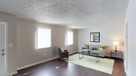 This two-floor space presents a welcoming and functional layout, highlighted by a generously sized living area that serves as the focal point of the main level. The living room features rich, dark hardwood flooring that contrasts attractively with the soft, neutral gray walls, creating a warm and contemporary atmosphere. A textured ceiling adds subtle visual interest overhead, while crisp white baseboards frame the room cleanly.

Two large windows fitted with vertical blinds line one wall, drawing in abundant natural light that brightens the entire space throughout the day. The open layout provides ample room for versatile furniture arrangements, as demonstrated by the combination of a gray sofa with accent pillows, a sleek glass-top coffee table, and an accent armchair positioned on a geometric-patterned area rug.

Wall space is thoughtfully utilized, with a framed artwork piece anchoring the seating area and adding a decorative touch. The overall ambiance of the living area is calm and neutral, offering a flexible foundation for a variety of interior styles. The two-floor configuration suggests additional living or sleeping quarters above, providing a practical separation between common and private areas of the home.