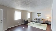 This two-floor space presents a welcoming and functional layout, highlighted by a generously sized living area that serves as the focal point of the main level. The living room features rich, dark hardwood flooring that contrasts attractively with the soft, neutral gray walls, creating a warm and contemporary atmosphere. A textured ceiling adds subtle visual interest overhead, while crisp white baseboards frame the room cleanly.

Two large windows fitted with vertical blinds line one wall, drawing in abundant natural light that brightens the entire space throughout the day. The open layout provides ample room for versatile furniture arrangements, as demonstrated by the combination of a gray sofa with accent pillows, a sleek glass-top coffee table, and an accent armchair positioned on a geometric-patterned area rug.

Wall space is thoughtfully utilized, with a framed artwork piece anchoring the seating area and adding a decorative touch. The overall ambiance of the living area is calm and neutral, offering a flexible foundation for a variety of interior styles. The two-floor configuration suggests additional living or sleeping quarters above, providing a practical separation between common and private areas of the home.