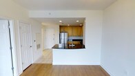 This single-floor space is designed with an open and functional layout that seamlessly connects the main living area to the kitchen. The kitchen is defined by warm honey-toned wood cabinetry that extends to the upper walls, providing ample storage. Dark granite countertops contrast neatly against the cabinetry, adding a polished aesthetic. A curved peninsula extends outward, creating a natural boundary between the kitchen and living area while offering additional counter space. Stainless steel appliances, including a full-sized refrigerator, contribute a modern, clean look to the culinary space. Recessed lighting is installed throughout the kitchen area, ensuring well-distributed illumination. The living area features warm hardwood flooring, while the kitchen and entryway transition to light-toned ceramic tile, providing a practical and visually distinct zoning effect. Crisp white walls throughout the space enhance the sense of openness and brightness. A white entry door with a floor-level vent is visible at the rear, indicating a well-integrated HVAC system. Overall, the space presents a clean, contemporary feel suited for everyday living.