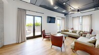 Located at 600 North Avenue in New Rochelle, NY, this single-floor commercial space offers a thoughtfully designed interior that blends industrial aesthetics with contemporary comfort. The open layout encompasses three distinct functional areas, all unified by warm light hardwood flooring, crisp white walls, and an exposed ceiling with visible ductwork and sleek linear lighting — hallmarks of modern industrial-chic design.

The lounge area serves as a welcoming social hub, furnished with plush neutral sofas, cognac leather accent chairs, and a glass coffee table. Bold black-and-white graphic wall art adds an artistic edge, while large glass doors open directly to an outdoor terrace, flooding the space with natural light and providing seamless indoor-outdoor connectivity.

A dedicated conference and dining area features a long white communal table surrounded by black chairs, complemented by a wall-mounted display screen. Sheer linen curtains frame multiple windows, softening the industrial ceiling elements while maintaining a bright, airy atmosphere.

The integrated kitchenette and service zone is outfitted with an expansive run of bold navy cabinetry, a white countertop with a built-in sink, and an additional wall-mounted screen — ideal for presentations or collaborative work. Together, these areas create a versatile, stylish environment well-suited for professional gatherings and creative collaboration.
