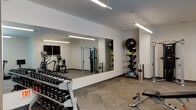 This modern fitness center is thoughtfully designed to support a comprehensive workout experience within a single-floor layout. The space features a clean, contemporary aesthetic with neutral white walls, herringbone-patterned carpet flooring, and bright linear pendant lighting that ensures excellent visibility throughout.

The cardio zone is anchored by multiple treadmills positioned in front of large windows, allowing natural light to fill the area and providing views of the surrounding exterior. An elliptical trainer and a stationary bike round out the cardiovascular equipment offerings. A wall-mounted flat-screen television is conveniently positioned nearby for entertainment during workouts.

The strength training area includes a full dumbbell rack with an extensive range of free weights, an adjustable weight bench, and a Hoist dual-function cable machine suitable for a wide variety of resistance exercises. A large floor-to-ceiling mirror spans one wall, visually expanding the space and allowing users to monitor their form.

The functional training section is equipped with medicine balls on a vertical storage rack, a BOSU balance trainer, and additional accessories for dynamic and core-focused exercises. The overall layout is open and well-organized, providing ample space to move freely between equipment zones, making this fitness center a practical and fully equipped facility for users of all fitness levels.