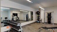 This modern fitness center is thoughtfully designed to support a comprehensive workout experience within a single-floor layout. The space features a clean, contemporary aesthetic with neutral white walls, herringbone-patterned carpet flooring, and bright linear pendant lighting that ensures excellent visibility throughout.

The cardio zone is anchored by multiple treadmills positioned in front of large windows, allowing natural light to fill the area and providing views of the surrounding exterior. An elliptical trainer and a stationary bike round out the cardiovascular equipment offerings. A wall-mounted flat-screen television is conveniently positioned nearby for entertainment during workouts.

The strength training area includes a full dumbbell rack with an extensive range of free weights, an adjustable weight bench, and a Hoist dual-function cable machine suitable for a wide variety of resistance exercises. A large floor-to-ceiling mirror spans one wall, visually expanding the space and allowing users to monitor their form.

The functional training section is equipped with medicine balls on a vertical storage rack, a BOSU balance trainer, and additional accessories for dynamic and core-focused exercises. The overall layout is open and well-organized, providing ample space to move freely between equipment zones, making this fitness center a practical and fully equipped facility for users of all fitness levels.