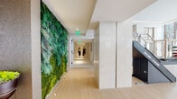 This single-floor wellness facility is thoughtfully designed to offer a comprehensive health and beauty experience. The entrance corridor immediately captures attention with a dramatic floor-to-ceiling living green wall, composed of rich layers of moss and ferns in varied shades of green. Light wood flooring runs throughout this hallway, complemented by smooth, stone-clad columns and recessed lighting that creates a polished, inviting atmosphere.

The fitness area is expansive and well-appointed, featuring an extensive array of professional-grade strength and cardio equipment. Large floor-to-ceiling windows flood the gym with natural light and frame views of the surrounding cityscape, while a coffered ceiling with integrated lighting adds architectural refinement overhead.

A dedicated body composition assessment room offers a clinical yet clean environment, housing a digital body scanning station for precise health metrics. The spa corridor presents a series of private treatment rooms, including a serene massage room with a cream-upholstered treatment table and soft neutral walls. A specialized wellness pod and glass-partitioned relaxation areas enhance the spa's therapeutic offerings.

The Glam Room, identified by frosted glass signage, provides a professional beauty treatment space complete with a styling chair and service station, rounding out this facility's full-service wellness offerings.