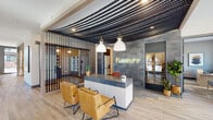 Located at 510 E Pettigrew St in Durham, NC, The Ramsey presents a thoughtfully designed amenity space that blends contemporary aesthetics with warm, inviting details. The entrance welcomes visitors through floor-to-ceiling glass doors into a bright, open lobby featuring light wood-look tile flooring, tall indoor greenery, and a distinct reception desk adorned with white tile and a bold branded sign. An eye-catching textured ceiling treatment and pendant lighting complete the arrival experience. Adjacent to the lobby, a comfortable lounge area showcases a patterned feature wall, a statement drum chandelier, built-in cabinetry with glass fronts, and plush seating arranged around a central coffee table — ideal for relaxing or informal gatherings. A cozy nook with teal upholstered banquette seating, marble-topped tables, and decorative pendant lights offers an intimate setting for smaller conversations or casual dining. The kitchen area features a sleek dark island with built-in appliances, patterned tile flooring, and a suspended open shelving unit lush with potted plants, adding a lively, organic touch. A private workspace with a dark desk and artistic wall décor provides a focused environment, while a sitting alcove with floor-to-ceiling windows brings in natural light and outdoor views, rounding out this well-appointed community space.