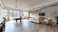 This expansive single-floor recreational space showcases a modern industrial design, blending warm neutral tones with rustic wood-look flooring that runs throughout. Soaring ceilings with exposed ductwork add an urban loft character, while a series of pendant lights provide focused illumination across the open floor plan.

The main gathering area is anchored by a large sectional sofa and accent chairs arranged around a wall-mounted flat-screen television set atop a sturdy industrial-style wood and metal media console. Artwork featuring equestrian themes lends a curated, artistic touch to the warm taupe walls. Adjacent to the lounge area, a full-size pool table serves as a centerpiece for recreation, positioned to take full advantage of the generous floor space.

A dining table with seating for several guests occupies one corner, ideal for group gatherings or casual meals. Floor-to-ceiling black-framed windows line one wall, flooding the interior with natural light and offering open views of the surrounding landscape.

A dedicated wet bar area features rich wood cabinetry, a stone tile backsplash, a sink, floating shelves, and bar stool seating — perfect for entertaining. A barn-style door adds a rustic design accent nearby. Together, these elements create a versatile, welcoming space well-suited for socializing and leisure.
