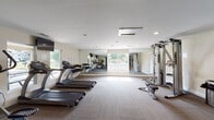 This single-level fitness center is thoughtfully designed to support a comprehensive workout experience. The main exercise area is bright and open, featuring large windows along multiple walls that invite abundant natural light and provide views of the surrounding outdoor greenery. Full-length mirrors are strategically placed to visually expand the space and allow exercisers to monitor their form.

The cardio zone is well-stocked with multiple True-brand treadmills and elliptical machines, including a recumbent cross-trainer, all arranged to take advantage of the natural light and outdoor views. A wall-mounted flat-screen television accompanies this area for entertainment during workouts. A multi-station cable weight machine and a dedicated free weights section, complete with a dumbbell rack and adjustable bench, round out the strength training offerings.

A separate functional training area features teal accent walls and houses a robust selection of equipment, including a suspension training rig, medicine balls, kettlebells, a punching bag, and stability balls on a storage stand. The flooring throughout consists of durable, multi-toned carpet tiles, and recessed lighting supplements the natural light. A glass-panel entry door provides easy outdoor access.