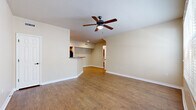 This single-floor residence offers a thoughtfully designed layout that balances open living spaces with comfortable private areas. The main living room is generously proportioned, featuring warm wood-look laminate flooring, neutral beige walls, and a ceiling fan with integrated lighting. A large picture window with horizontal blinds fills the space with natural light and provides views of mature trees and greenery outside. The living area connects seamlessly to the kitchen through a pass-through bar counter, which offers a practical and social layout for daily living. The kitchen is equipped with warm wood cabinetry, dark granite-style countertops, track lighting, and black appliances including a refrigerator and range. A dedicated utility panel is conveniently accessible within the kitchen corridor. The bedrooms are carpeted for comfort, each featuring ceiling fans, natural light from windows, and neutral tones that create a calm atmosphere. The bathroom is well-appointed with a single vanity topped with granite-style counters, warm wood cabinetry with storage above, a large mirror, and a full tub-shower combination with white tile surround. Throughout the home, clean white baseboards and consistent neutral tones create a cohesive, move-in ready aesthetic.