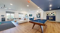 Located at 14300 Boggy Creek Rd in Orlando, FL, this generously sized single-floor community amenity space is thoughtfully designed for recreation and social gatherings. The open-plan layout is anchored by warm light wood flooring, crisp white walls, and a striking navy blue ceiling accent that adds a bold, modern touch throughout.

The main lounge area features a large sectional sofa with colorful accent pillows, curated wall art, and a wall-mounted flat-screen TV, creating an inviting space to relax. Adjacent to the lounge is a stylish wet bar area with teal cabinetry, white countertops, a sink, and built-in microwave — ideal for entertaining. A pair of blush accent chairs and a bold abstract painting complete this vibe.

The game zone includes a full-size pool table and an air hockey table, illuminated by contemporary sputnik-style pendant chandeliers. Large floor-to-ceiling sliding glass doors flood this area with natural light and open onto an outdoor terrace with seating overlooking lush greenery.

A dedicated sports simulator room features a large projection screen with a golf and multi-sport simulation setup, rubber sport flooring, a green turf mat, and a monitor station — offering an immersive athletic experience. Additional café-style seating and high-top bar stools provide flexible gathering spots throughout the space.