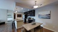 This single-floor space showcases an open-concept layout that seamlessly integrates the kitchen and dining areas into one cohesive living environment. The kitchen is appointed with floor-to-ceiling dark cabinetry that provides ample storage, offering a bold contrast against the neutral gray walls. Stainless steel appliances, including a French door refrigerator, electric range, and built-in microwave, lend a contemporary, functional edge to the space. A peninsula with granite countertops and bar seating extends from the kitchen, doubling as a casual dining and food prep area, complete with an undermount sink and modern faucet. Recessed lighting runs throughout the ceiling, supplemented by elegant pendant lights that hang above both the peninsula and dining area, creating layered illumination. The dining space accommodates a light wood table with coordinating seating, grounded by rich dark hardwood flooring that flows throughout. A framed chalkboard near the entryway adds a personalized, welcoming touch, while a large canvas artwork on the dining wall enhances the modern aesthetic. The overall ambiance is contemporary and inviting.
