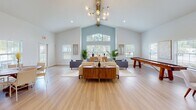 Located at 2335 Downing Ave in Savannah, Georgia, this single-floor community space offers an inviting and well-appointed environment defined by its striking architectural character and modern design sensibility. The centerpiece of the space is a grand open living area anchored by soaring vaulted ceilings with an exposed wooden beam and an elegant multi-globe chandelier that draws the eye upward. A dramatic arched window wall floods the room with natural light, complemented by additional windows on all sides that frame views of the surrounding greenery. Light hardwood flooring flows seamlessly throughout, reinforcing the airy, cohesive aesthetic. The main lounge area features contemporary mid-century inspired furnishings, including plush seating arrangements and warm wood accent pieces, creating a comfortable and social atmosphere. A shuffleboard table adds a recreational element to the space. Adjacent to the lounge, a dedicated dining area provides seating for group gatherings, with direct access to an exterior door. The fully equipped kitchen is finished with slate-gray shaker cabinetry, white quartz countertops, a tile backsplash, stainless steel appliances, and a large center island, making it ideal for hosting events. A secondary prep or beverage station offers additional counter space and small appliance hookups. Together, these areas create a versatile, welcoming community hub suited for a wide range of gatherings and activities.