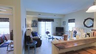 This single-floor space offers a warm and inviting atmosphere with a thoughtfully arranged living area at its heart. The living room is generously sized, featuring plush carpeted flooring that adds a sense of comfort underfoot. A large sliding glass door floods the room with natural light and provides a view to an outdoor patio or balcony, creating a bright and airy ambiance. Dark curtain valances frame the windows and glass door, adding a touch of contrast to the soft, neutral-toned walls.

The living room is furnished with a relaxed seating arrangement, including a cushioned sofa and a classic rocking chair, centered around a traditional coffee table. A flat-screen television is positioned on a media stand, making this an ideal space for relaxation and entertainment. Decorative accents, including an ornate blue display cabinet, framed wall art, and charming figurines, lend the space a personalized and homey character.

An open-concept transition connects the living room to a dining or kitchen counter area, which features a rustic wooden surface, hinting at a seamlessly flowing layout. Ceiling vents suggest a central HVAC system, ensuring year-round comfort. Overall, the space presents a lived-in, welcoming environment with functional design and distinctive personal touches throughout.