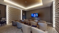 This private screening room is designed to deliver a premium cinematic experience within an elegantly appointed setting. The room accommodates multiple rows of plush, curved lounge chairs upholstered in neutral tones, each accompanied by a small side tray, ensuring comfort during extended viewing sessions. Seating is arranged in a tiered layout to provide clear sightlines to the large wall-mounted flat-screen display positioned at the front of the room.

A defining architectural feature is the floor-to-ceiling three-dimensional geometric accent wall, finished in a warm woven diamond pattern and backlit to create a dramatic focal point. Dark taupe walls and recessed cove lighting along the tray ceiling contribute to an intimate, theater-like ambiance throughout the space.

Along one side of the room, a dark navy credenza provides storage and display space, accessorized with decorative objects and a colorful piece of wall art, adding a refined residential touch. A classic popcorn cart stationed near the accent wall reinforces the dedicated entertainment purpose of the space. Textured carpet flooring completes the room, enhancing acoustics and adding visual warmth to this thoughtfully designed screening lounge.