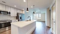 Located in the heart of Charlotte, NC, this contemporary single-floor residence offers a thoughtfully designed open-concept layout bathed in natural light. The kitchen serves as a central hub, anchored by a large white quartz waterfall island with an undermount sink and matte black faucet. Light wood-grain cabinetry extends to the ceiling, complemented by a horizontal tile backsplash and a full suite of stainless steel appliances, including a French door refrigerator, range, and built-in microwave. Stylish pendant lights hang above the island, adding a modern finishing touch. Hardwood-style flooring flows seamlessly throughout the living areas, creating a warm, cohesive aesthetic. The living space is generously proportioned, with large windows and a sliding glass door opening onto a private balcony, framing impressive views of the Charlotte skyline. A ceiling fan enhances comfort in the bright, airy bedroom, which also features a wide window overlooking the cityscape. Additional rooms provide flexible living space with neutral walls and consistent flooring. The bathroom features a white quartz vanity, light wood cabinetry with drawer storage, and a framed mirror. Recessed lighting throughout ensures a clean, well-lit environment across all areas of the residence.