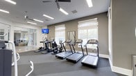 This single-floor fitness center offers a thoughtfully designed workout space that balances functionality with a clean, contemporary aesthetic. The room is finished with neutral taupe walls and durable dark carpeted flooring, creating a professional yet welcoming atmosphere. Large windows fitted with white roller shades line one wall, allowing generous natural light to fill the space while maintaining adjustable privacy. Overhead, modern linear LED lighting and a ceiling fan ensure consistent illumination and comfortable air circulation throughout workouts. The cardio equipment area is well-appointed, featuring a row of treadmills positioned along the windowed wall, complemented by elliptical trainers and additional cardio machines arranged behind them. Each piece of equipment includes an integrated digital display screen, providing users with entertainment and performance tracking capabilities. A wall-mounted flat-screen television is also present, offering an additional source of entertainment. Glass doors along one side of the room provide a bright, open connection to adjacent areas. The overall layout is spacious and uncluttered, reflecting a purposeful design that prioritizes ease of movement and a motivating environment for exercise.