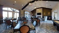 This single-floor communal space exudes a refined, welcoming ambiance designed for both social gatherings and relaxed lounging. The room's most striking architectural feature is its soaring vaulted ceiling, accentuated by bold dark wood beams that add warmth and character to the open interior. Geometric pendant lanterns hang at varying heights, casting a soft, inviting glow throughout the space and complementing the room's upscale aesthetic.

Large arched and floor-to-ceiling windows line the walls, flooding the interior with generous natural light and offering views of the surrounding landscape. The dining area is furnished with dark wood rectangular tables paired with stylish upholstered chairs featuring a distinctive orange geometric pattern, adding a vibrant pop of color against the neutral palette.

Toward the rear of the space, a cozy seating area is anchored by a fireplace set into a richly textured dark accent wall, with a wall-mounted television above and flanking sconce lighting. Plush wing chairs and accent seating create an intimate gathering nook. The entire space is unified by a sophisticated patterned area rug in cool blue tones. Together, these elements create a polished, multi-functional environment ideal for dining, conversation, and relaxation.